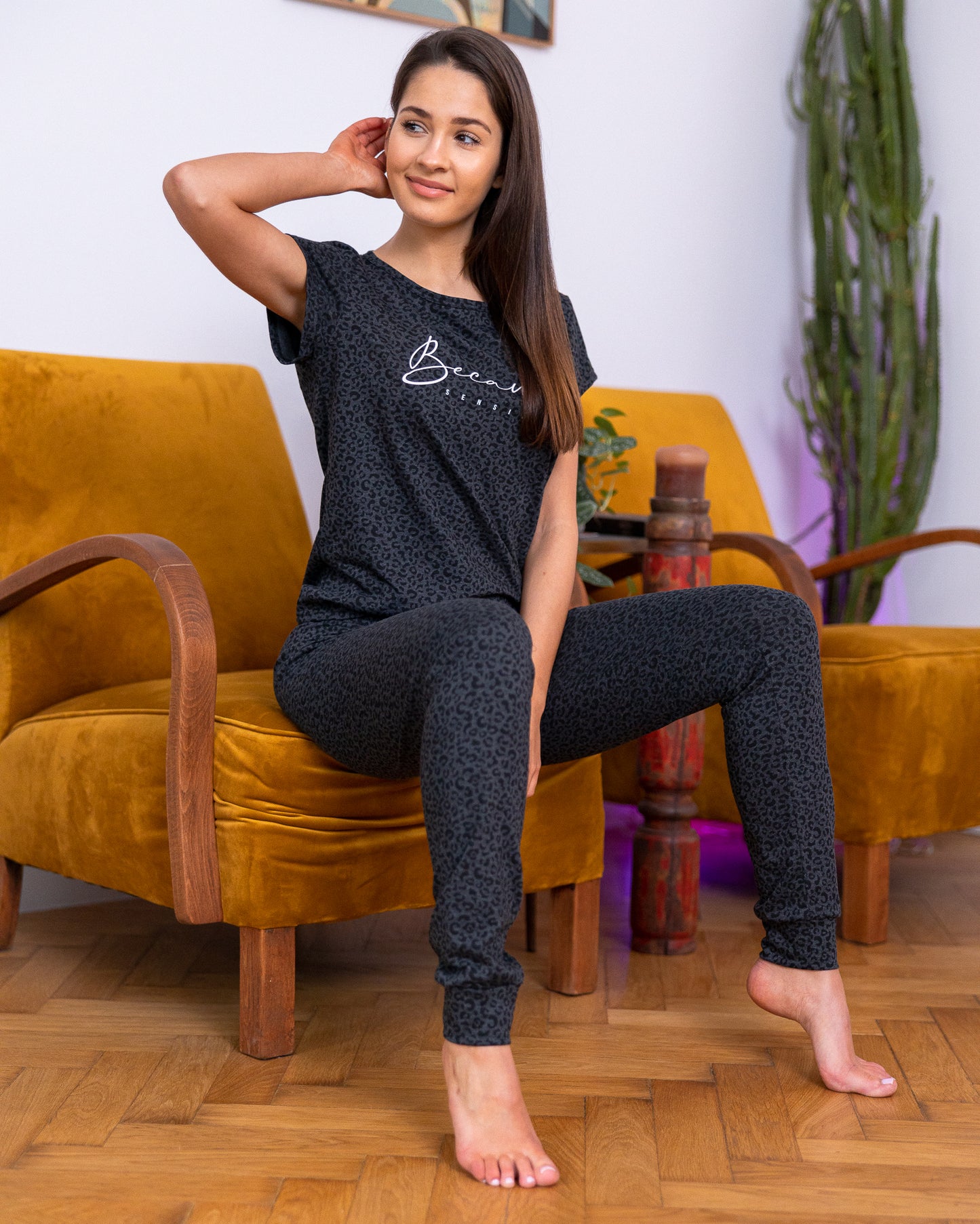 A Brunette female sitting on mustard suede loveseat wearing Two piece outfit set  Cotton-Blend Short Sleeve Top  Leopard Print Pattern  Soft Casual T-shirt  Long Pant Jogger  Cuff Ankles  Adjustable Satin Bow Tie  Sustainable pyjama set Eco-conscious lounge Wear 