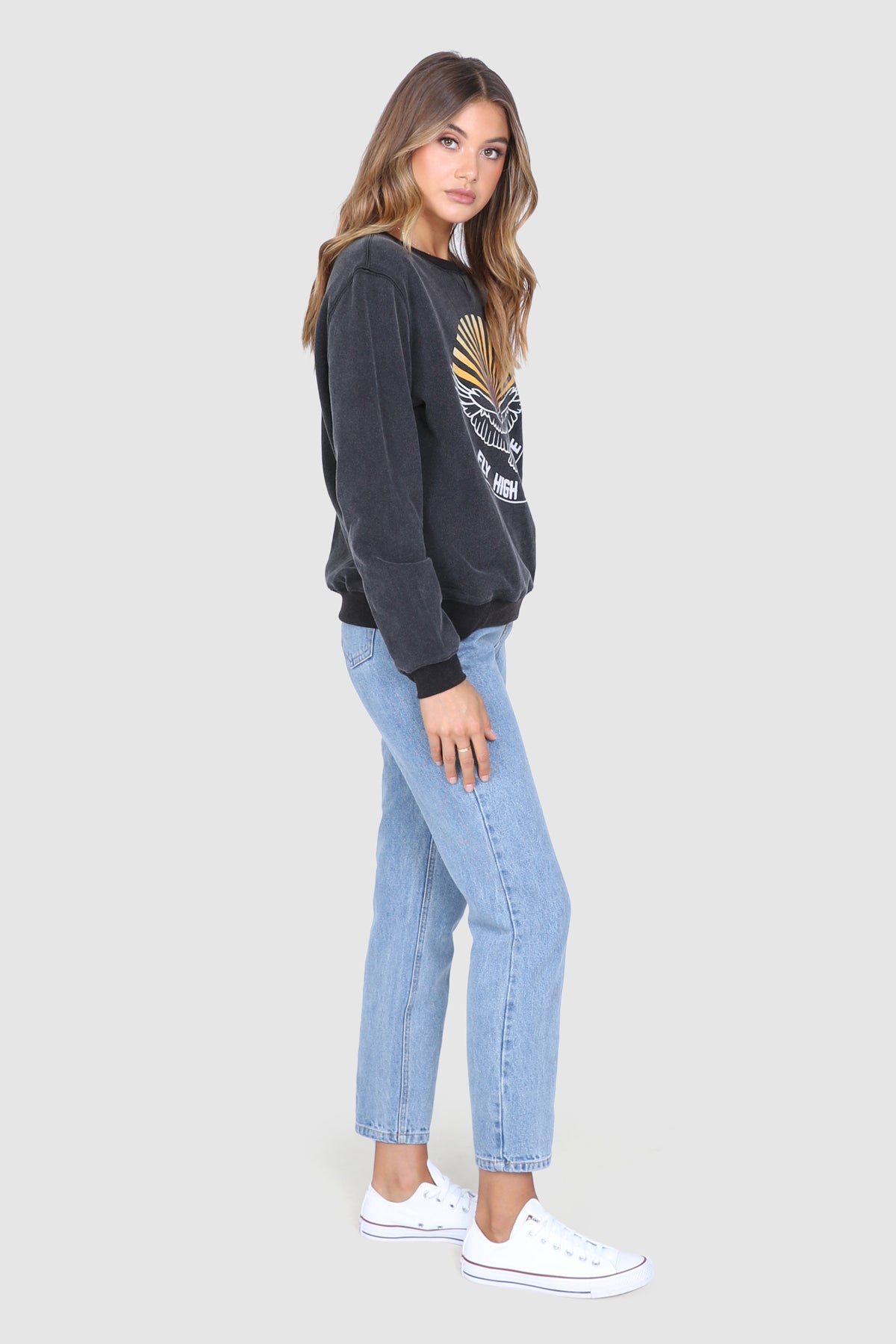 Bailage haired Caucasian Female wearing a  Vintage  Cotton Crew Neck  Round Neck  Eagle Graphic  Cuffed Long Sleeves  Relaxed Fit  Smoky Pullover paired with light washed mom jeans and white converse