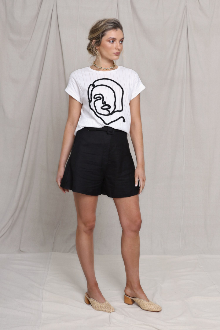 Blonde Caucasian Female wearing a Woman Face Line Art Graphic Tee  Ribbed Detail  Slightly Boxy-Fit  Scoop Neckline  White Short Sleeve T-shirt paired with a box cut linen black shorts and round toe woven beige mules