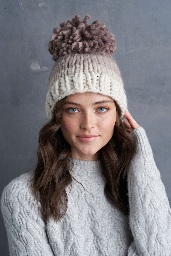 A Blue Eyed Brunette Caucasian female wearing a Chunky  Cable Knit  Striped  Beige undertone  Maroon Pom-Pom  Multicolored  Beanie paired with a turtleneck ribbed grey pullover sweater 