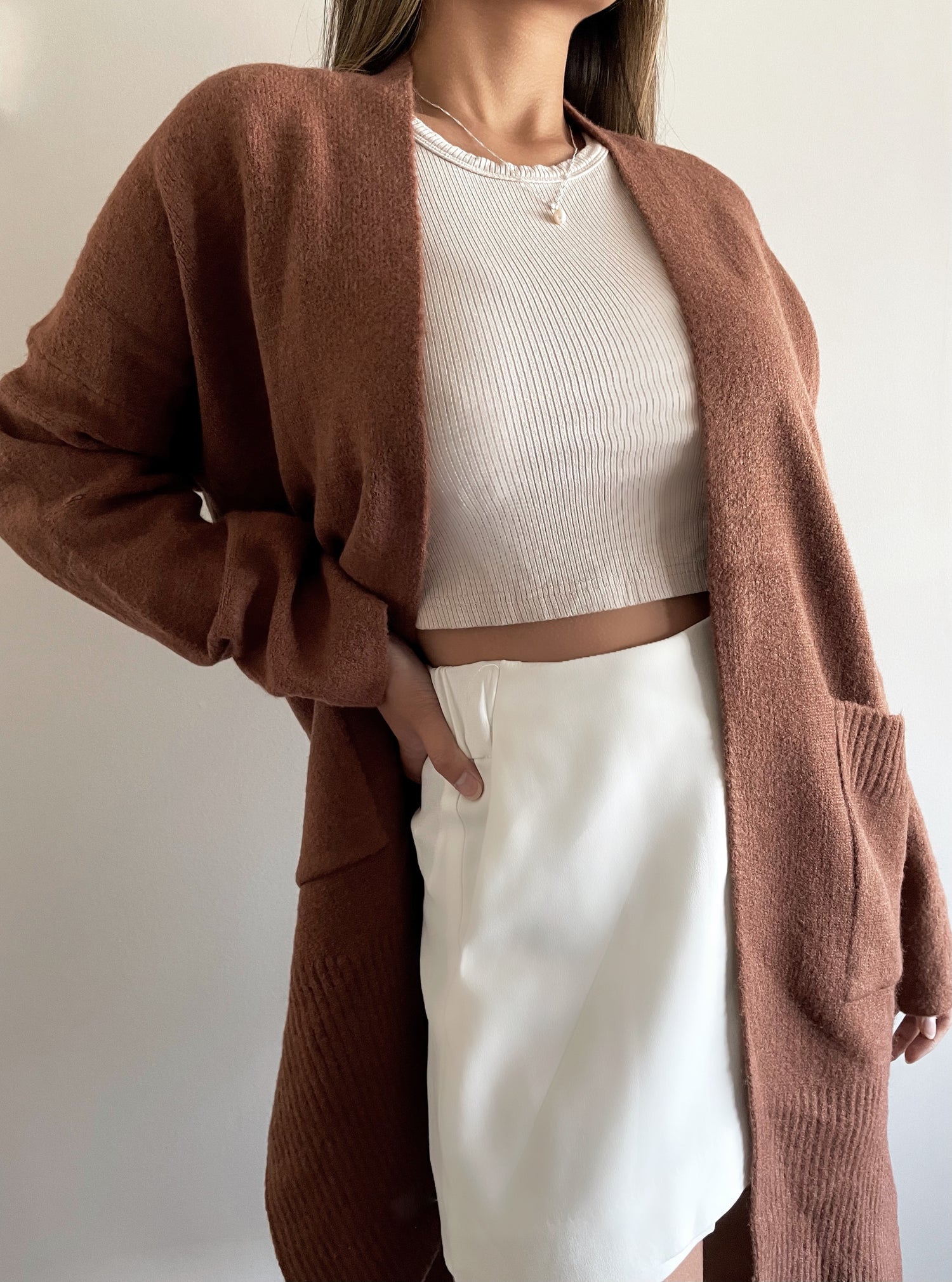 Asian Canadian female wearing wearing an Open Front Basic Versatile Dropped Shoulder Two Deep Side Pockets Two Side Split Slightly Oversized Ribbed Hemline And Cuffs Long Sleeved Cardigan in Rich Dark Chocolate paired with high scoop neck ribbed tank and white high waisted skirt 
