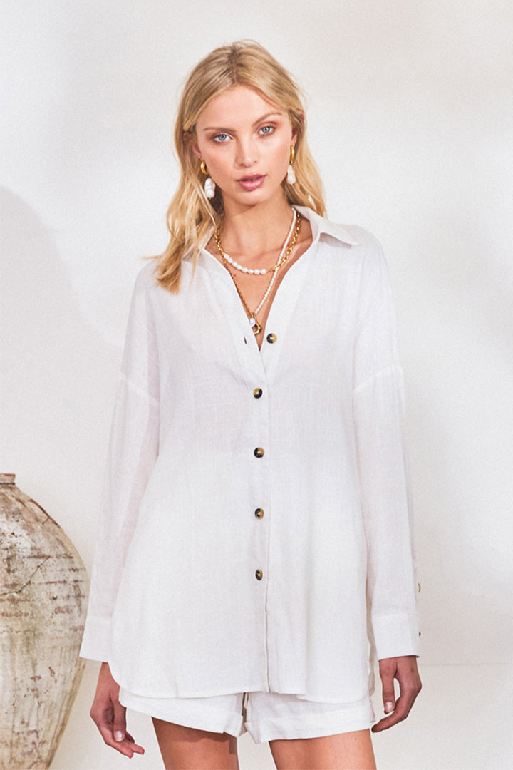 A blonde haired Caucasian female wearing a V neckline linen shirt Tortoiseshell buttoned down Side seam splits at hem Relaxed oversized fit collared blouse in ivory paired with white linen shorts