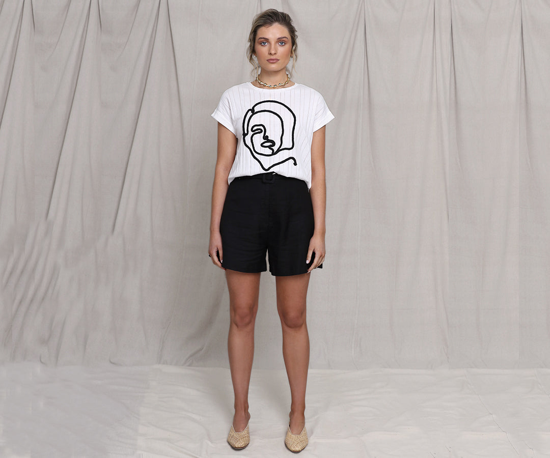 Blonde Caucasian Female wearing a Woman Face Line Art Graphic Tee Ribbed Detail Slightly Boxy-Fit Scoop Neckline White Short Sleeve T-shirt paired with a box cut linen black shorts and round toe woven beige mules
