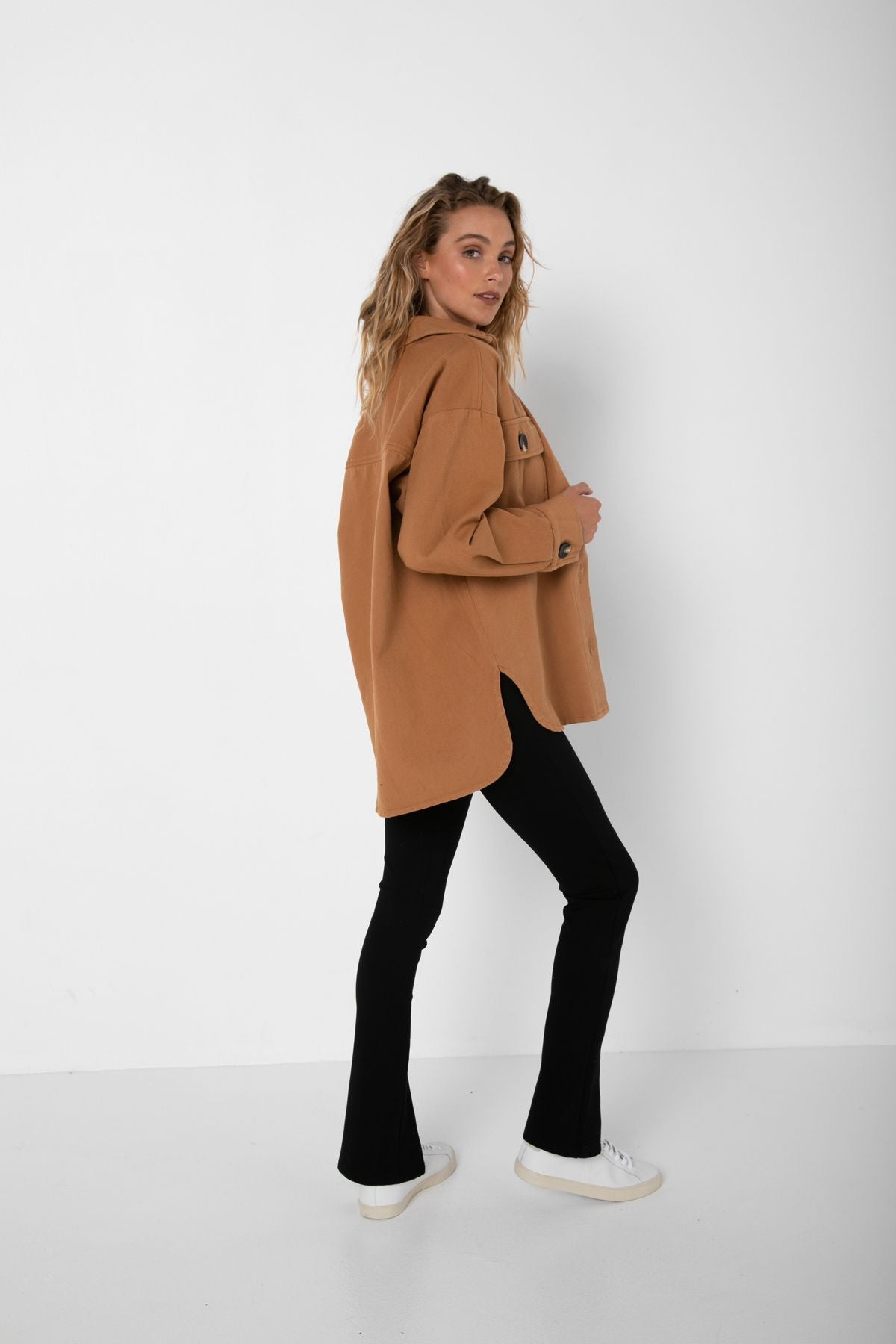 Curled Bailage Hair Caucasian Female wearing Long Sleeve Buttoned Down Top Lightweight Boyfriend Relaxed Fit Jacket Blouse Stylish Shacket Timeless Autumn and Winter Accessory Two Front Buttoned Pockets Side Slits in Tan paired with scoop neck black tee and Black high waisted stretchy long front slit pants and white sneakers